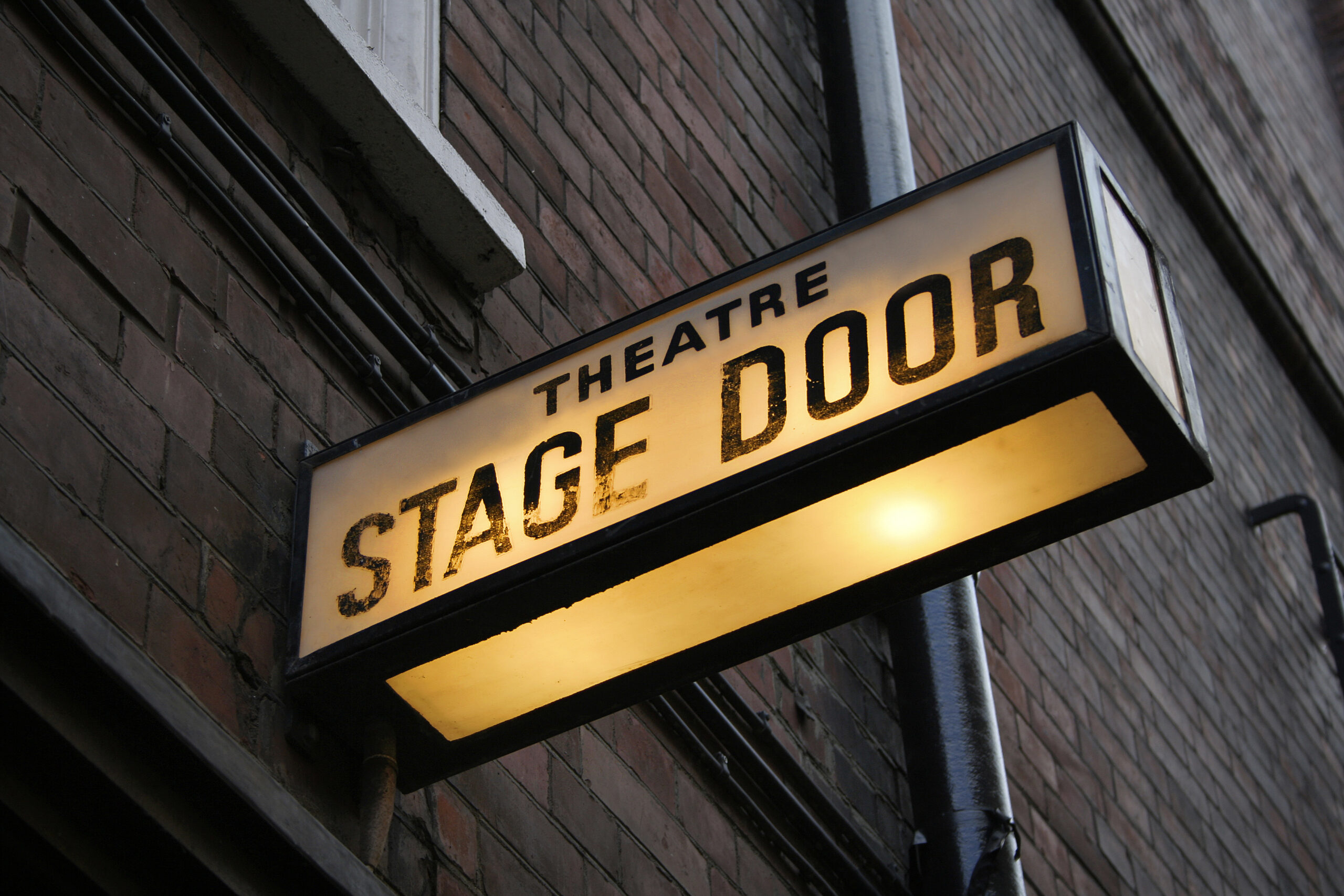 Close up photo of a back lit sign that reads "Theatre Stage Door"
